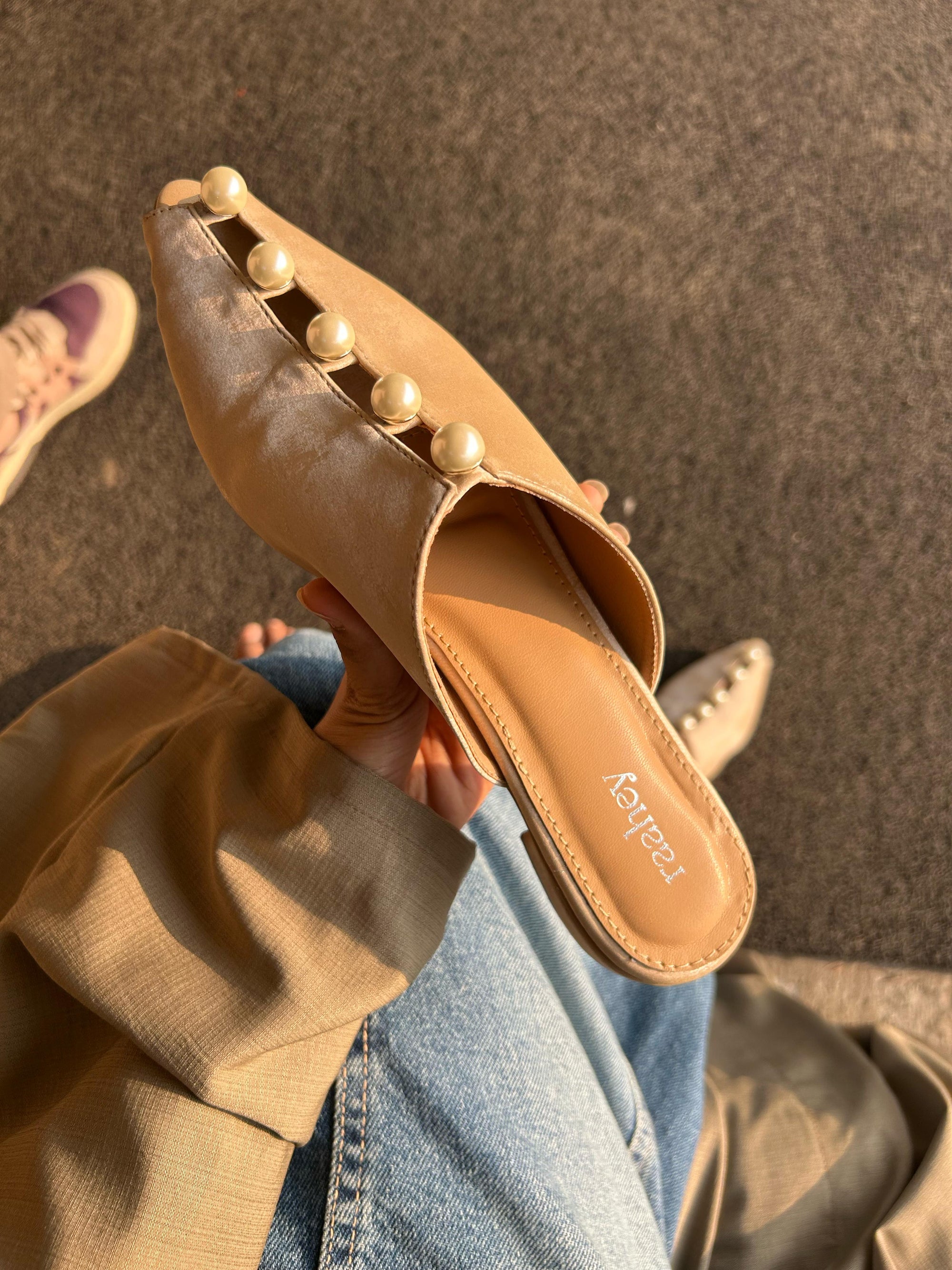 A person wearing beige mules with pearl embellishments, sitting in a vehicle.